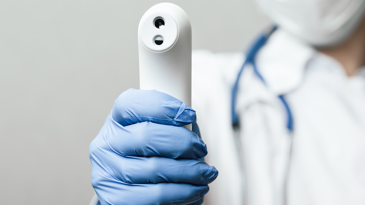 Nahaufnahme eines Arztes, der eine chirurgische Schutzmaske trägt, die bereit ist, das Infrarot-Stirn-Thermometer (Thermometer-Pistole) zu verwenden, um die Körpertemperatur auf Virussymptome zu überprüfen - Epidemic-Virus-Ausbruch-Konzept