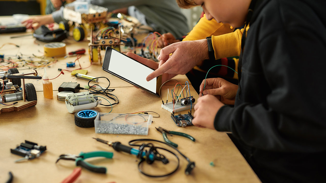 Aufschnitt von jungen Technikern, die mit einem Verkabelungskit in einer Robotikklasse arbeiten. Männlicher Lehrer hilft ihnen, Ratschläge geben, mit Tablet PC. Basteln, Bildungskonzept. Web-Banner