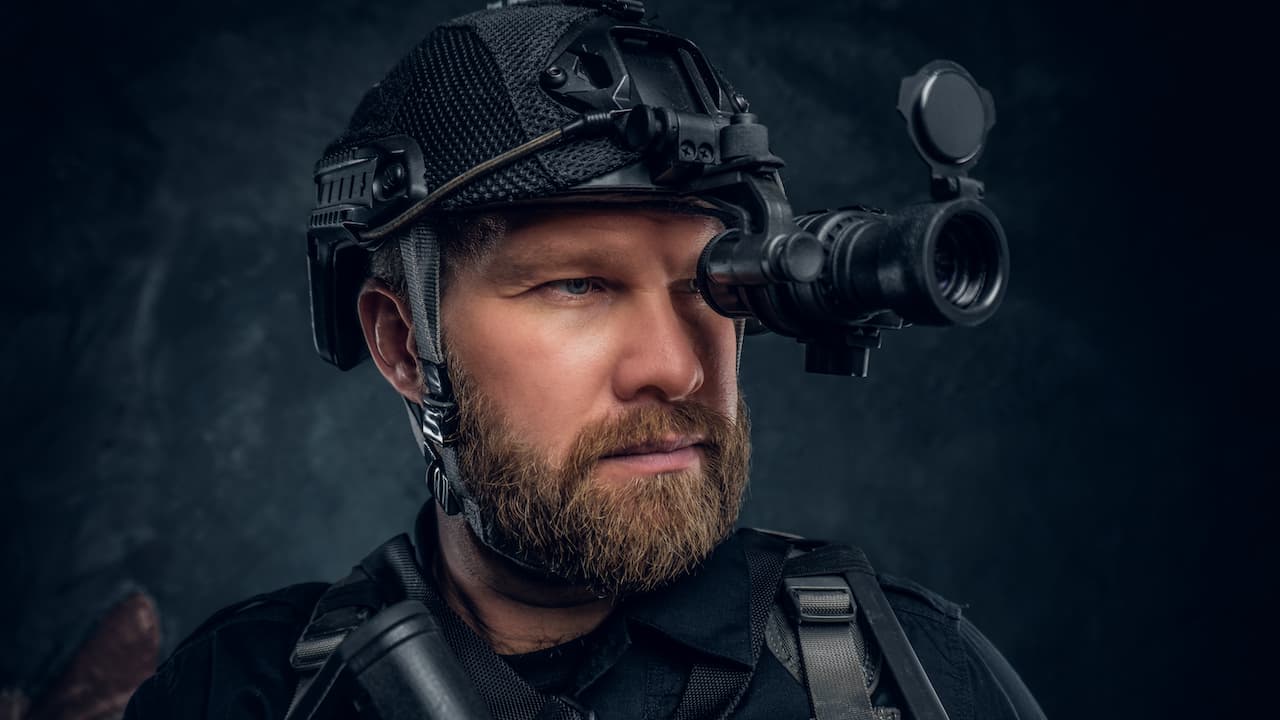 Close-up portrait of a bearded special forces soldier observes the surroundings in night vision goggles. Studio photo against a dark textured wall